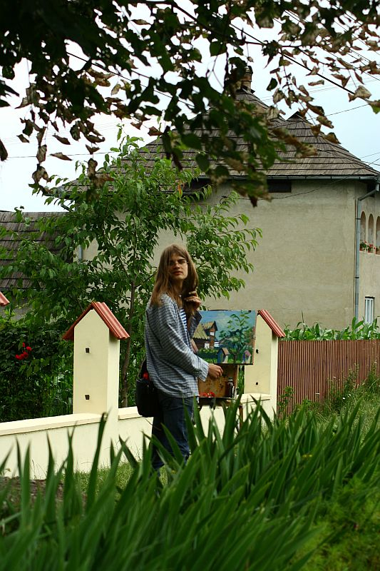 A woman stands by a white fence with birdhouses, holding a painting, amidst lush greenery with a residential home in the background in Romania. She seems to be either placing the artwork or admiring