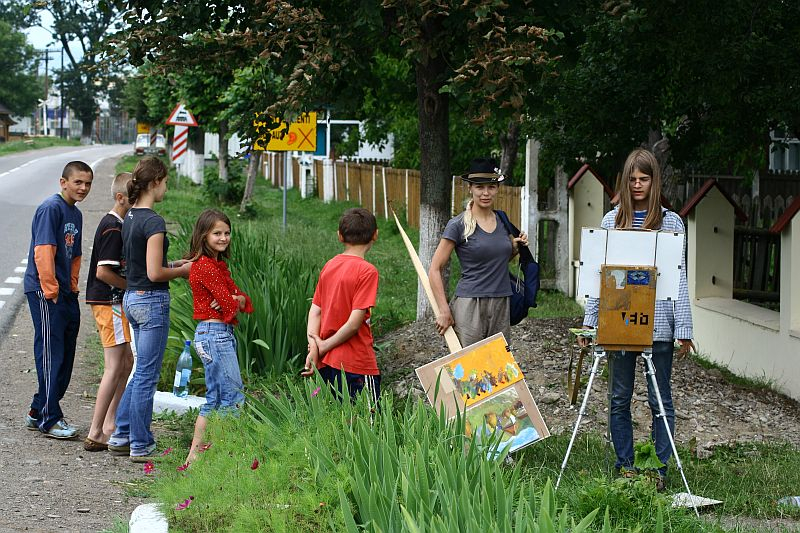 A group of children stand and watch a young artist painting outdoors by a roadside in Romania. The artist, wearing glasses and a hat, works on a landscape on an easel. They stand near a grassy area with a wooden fence and street signs in the background. The scene is relaxed, inviting travelers to enjoy local art tourism.