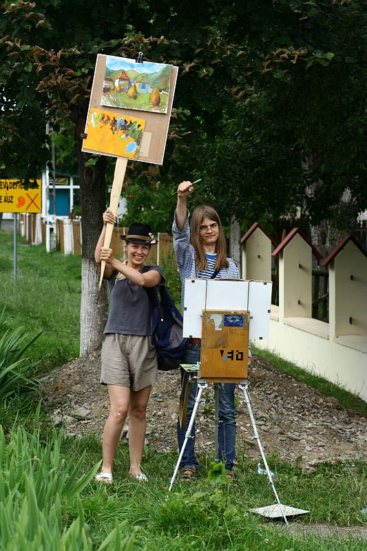 Two smiling artists in Romania outdoors with easels, one standing painting on a canvas, while the other showcases finished artwork.