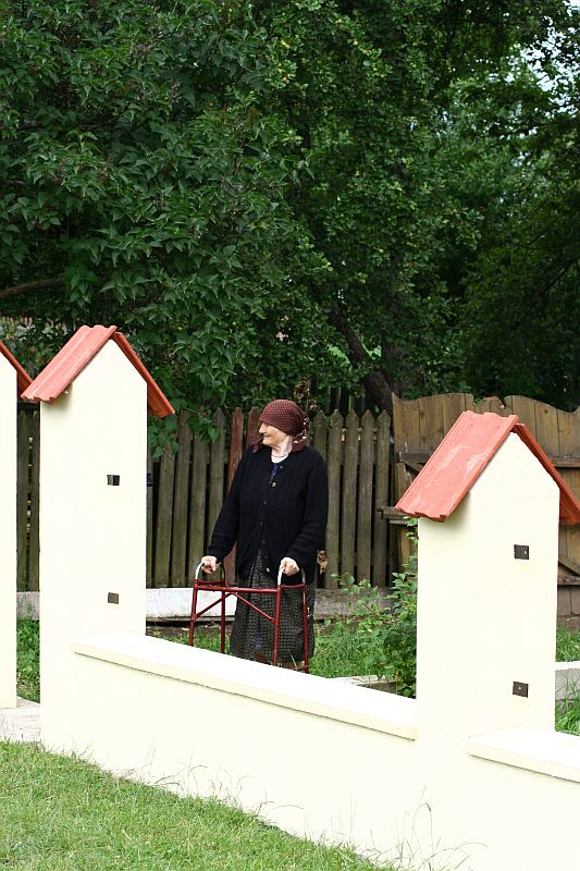 A person stands between two miniature houses, holding onto a red and white barrier, surrounded by lush greenery in what appears to be a park or outdoor exhibit in Romania, evoking a whimsical,