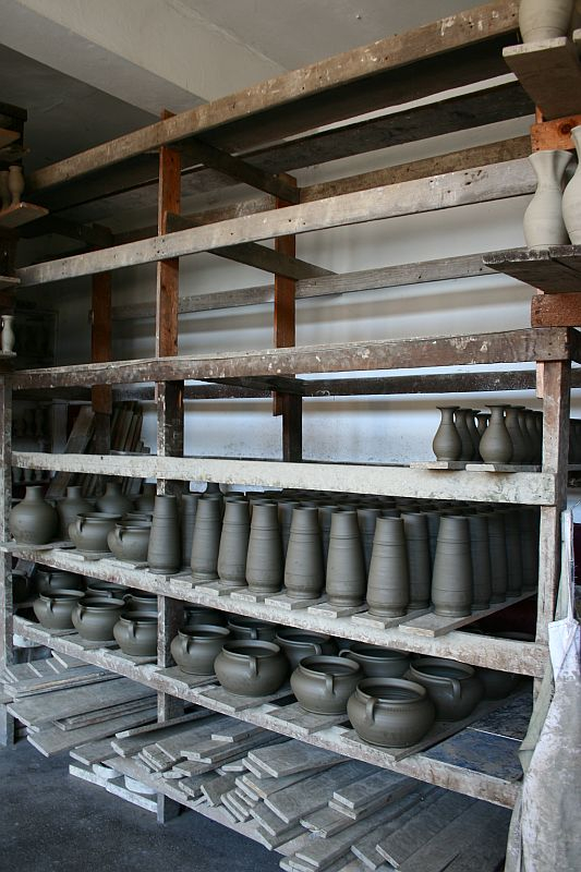 Rows of unfinished pottery pieces, including vases and bowls, sit on wooden shelves to dry in a Romanian workshop environment, showcasing various stages of the ceramic process.