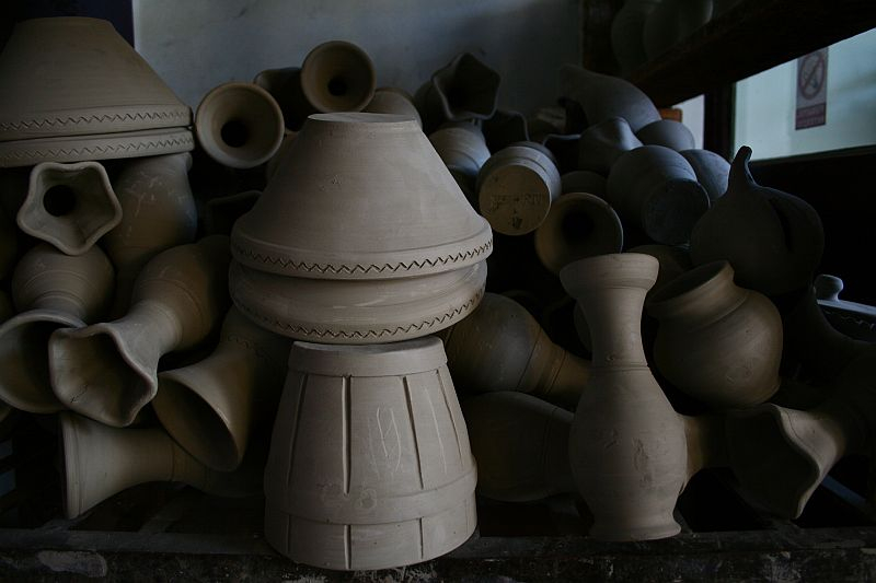 An assortment of unfinished clay pottery pieces, including pots and pitchers from Romania, set to dry, with prominent shadows casting a serene atmosphere over the traditional craft display.