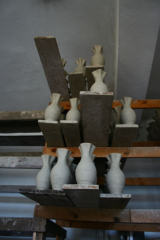 Unfinished ceramic vases at various stages of completion sit atop a wooden shelving unit in a Romanian pottery workshop, awaiting the next steps of their artistic transformation.