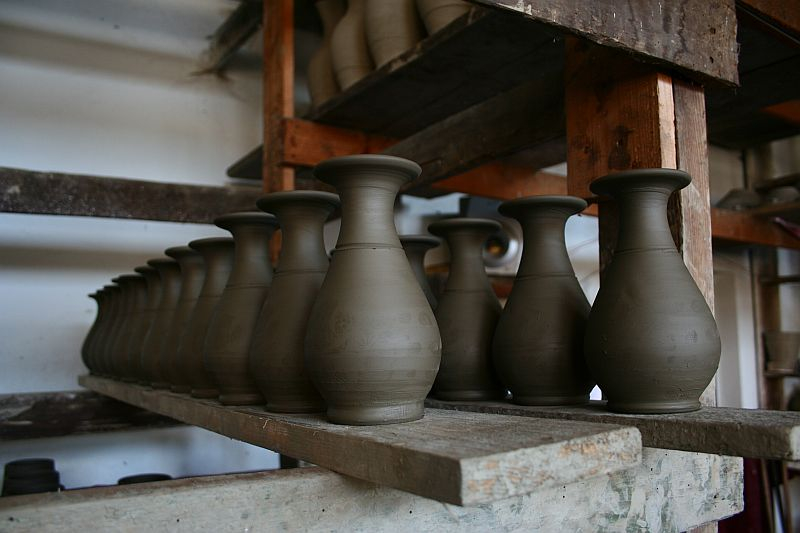 A row of unfinished clay pots lines up on a wooden shelf in a potter's workshop in Romania, waiting to be fired and glazed.