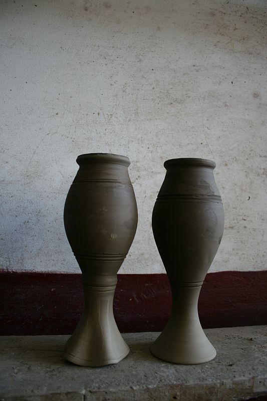 Two unfinished clay pots from Romania stand side by side against a white wall with a red baseboard, showcasing the symmetry and natural beauty of handcrafted pottery.