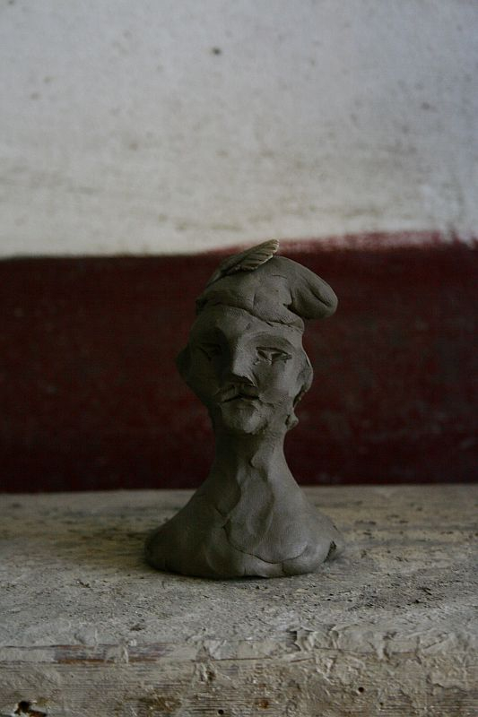 A handcrafted clay bust with distinct facial features sits on a wooden surface against a wall with peeling paint in Romania, accompanied by a perched butterfly on its head.