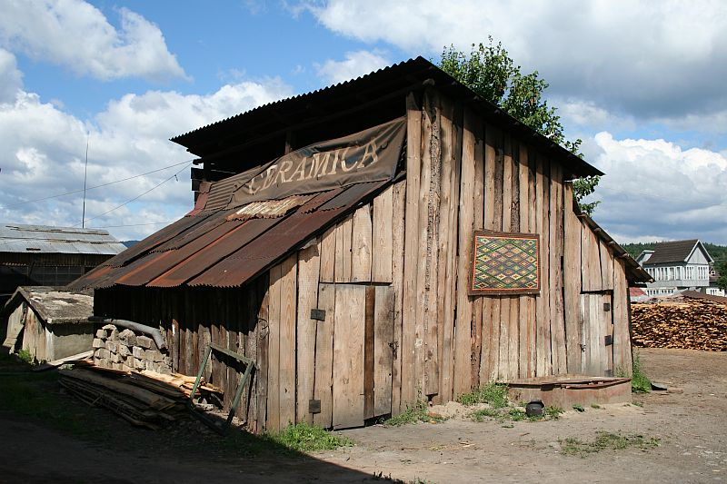 Rustic wooden shed with a corrugated iron roof appearing to sell drinks, adorned with a colorful geometric wall decoration, situated under a cloudy sky in Romania, with stacks of wood and residential buildings