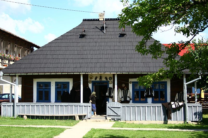 Traditional rural house in Romania with a symmetrical facade, featuring a blue wooden fence, ornate window frames, and a stone pathway leading to the entrance, set against a landscape with lush greenery and