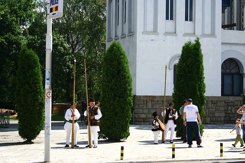 Individuals in traditional attire stand near a street sign in Bucharest, engaging in conversation, while others walk by on a sunny day with greenery and a white building in the background.