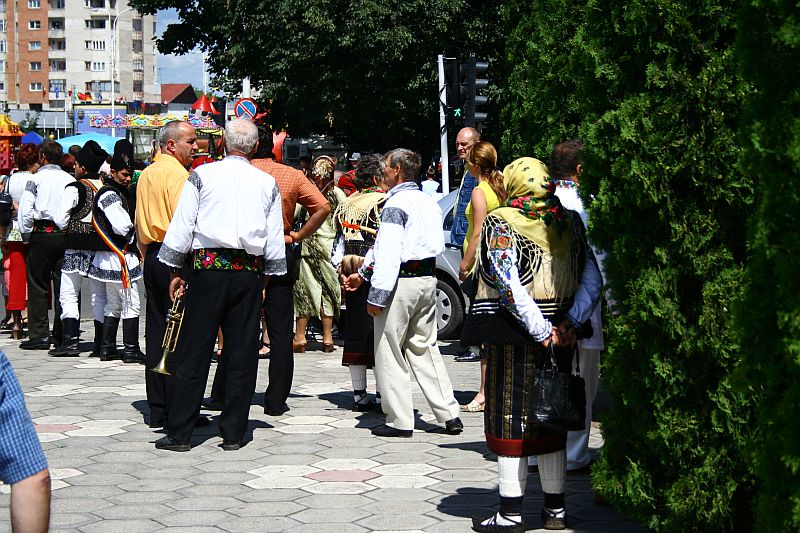 People in traditional Romanian attire gather on a sunny day, possibly for a cultural event or festival, marked by a convivial atmosphere amidst the urban surroundings.