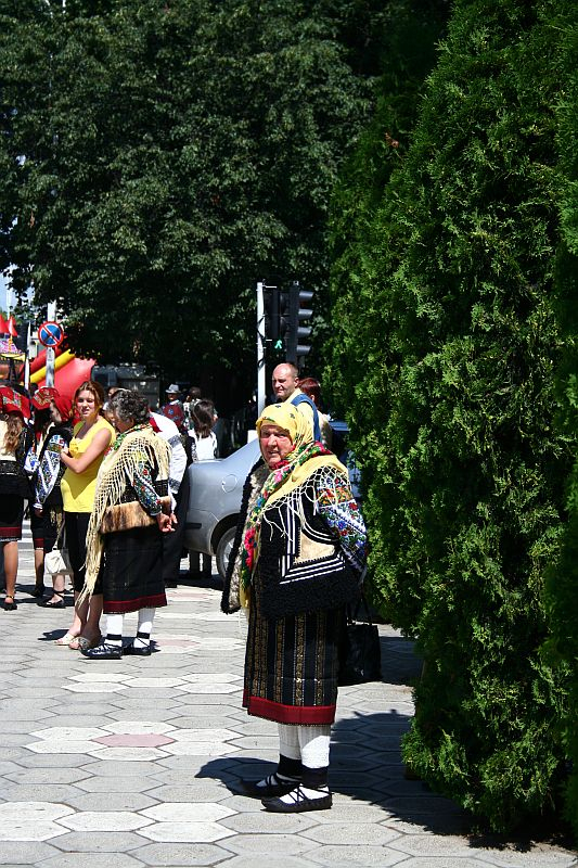 A woman dressed in traditional Romanian folk attire, including a patterned shawl and skirt, stands on a sunny street, with trees and a festive atmosphere in the background.