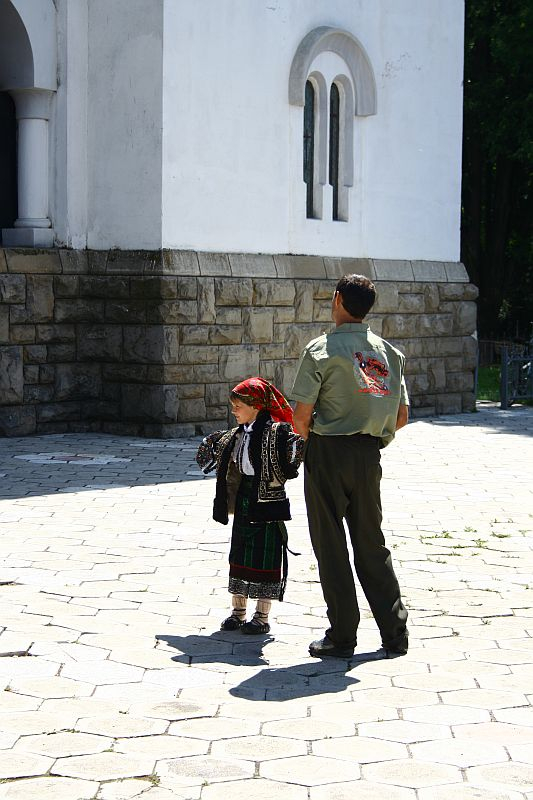 A child dressed in traditional Romanian folk attire walks alongside an adult in a military-style uniform, possibly during a cultural event or ceremony, in a courtyard against the backdrop of a white building with arched windows