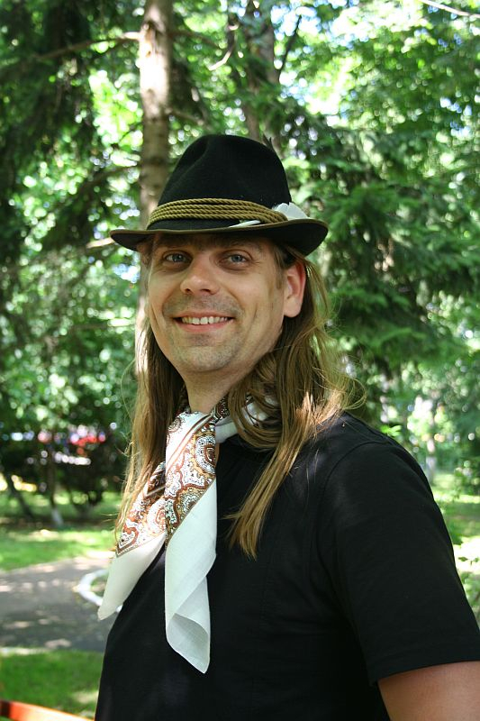 A smiling man with long hair wearing a black t-shirt, a patterned scarf, and a fedora hat poses casually in a lush, green park setting in Romania.