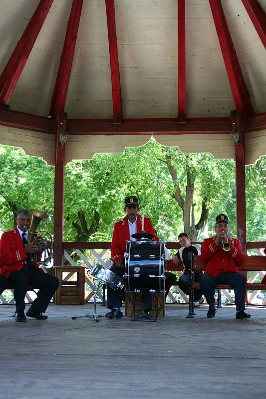 A quartet of musicians dressed in red and black perform under a gazebo in Romania, with trees in the background, featuring a trombone, drums, accordion, and trumpet.