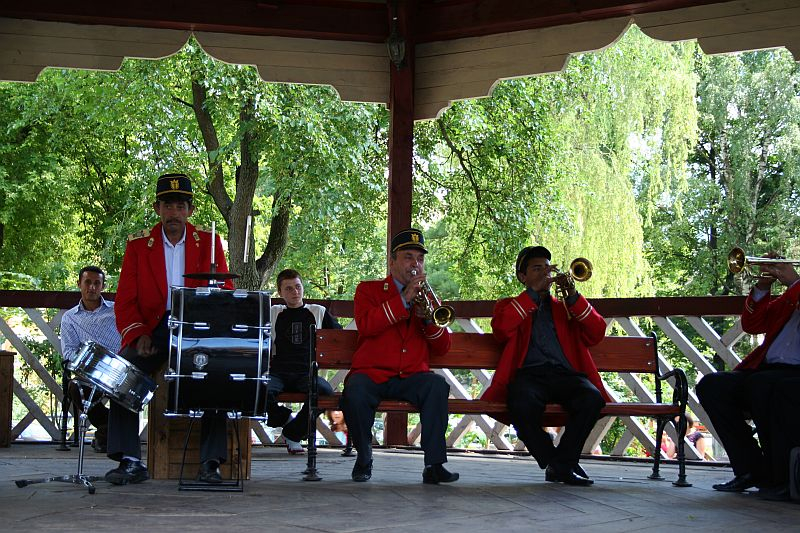 A brass band, dressed in traditional uniforms with red jackets and black hats, enthusiastically performs on a wooden gazebo amidst lush greenery in Romania, with a percussionist keeping the rhythm on a snare