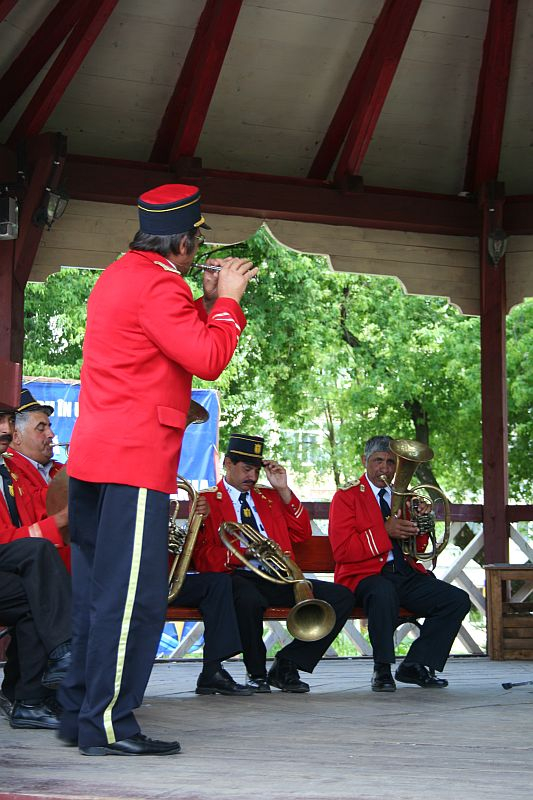 A band in vibrant red uniforms performs at an outdoor pavilion in Romania, with a focus on a standing trumpet player while his seated bandmates play brass instruments in the background, surrounded by lush greenery