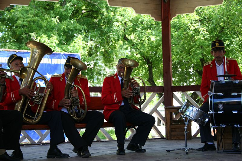 A brass band in red uniforms performs on an outdoor gazebo stage in Romania, with a tuba and horns in the foreground and a drummer at the back, all concentrating on their music.
