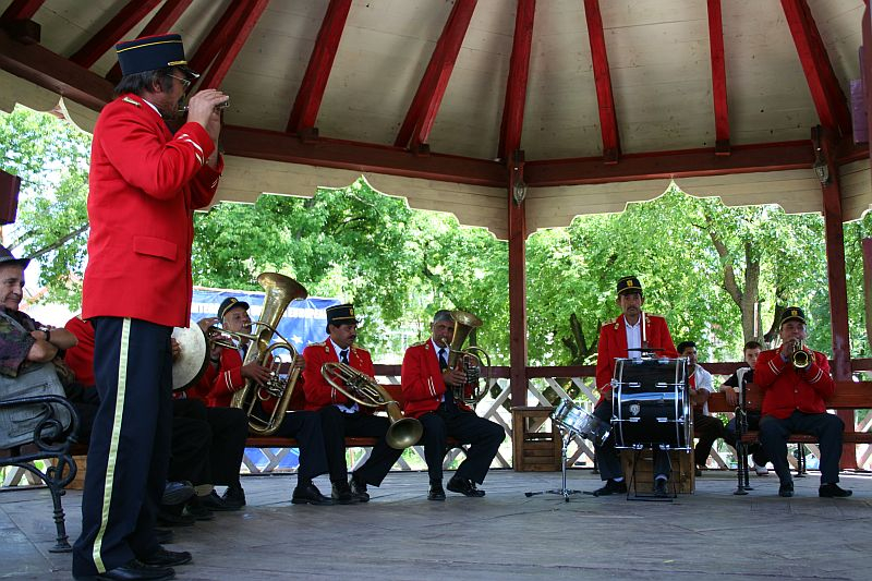 A brass band clad in traditional red and black uniforms performs enthusiastically under a gazebo in a lush, green park in Romania, with the conductor in front leading the ensemble.