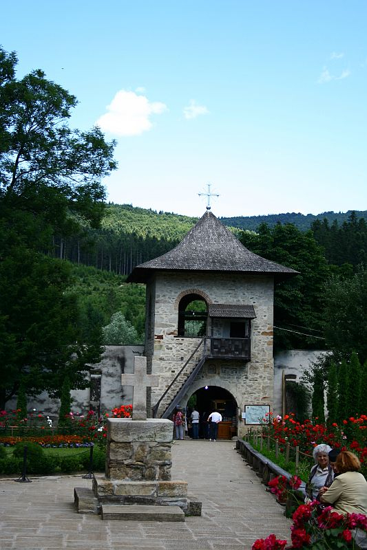 A rustic stone gateway with a wooden shingle roof leads into a serene courtyard surrounded by lush greenery in Romania, with visitors enjoying the tranquil surroundings.