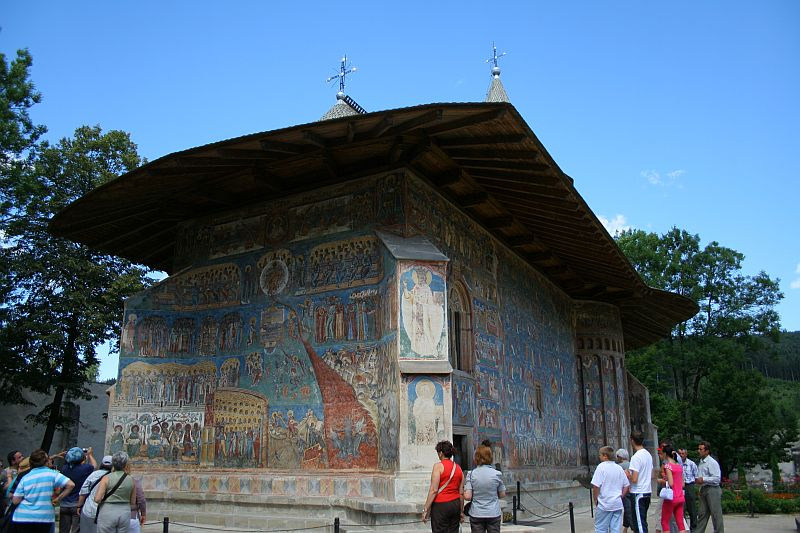 Visitors admire a historic painted monastery in Romania with intricate frescoes on its exterior walls, showcasing religious scenes under a clear blue sky.