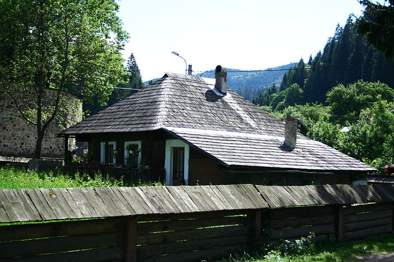 Traditional Romanian wooden house with a shingled roof, nestled in lush greenery under a clear blue sky, showcasing rural architecture and tranquility.