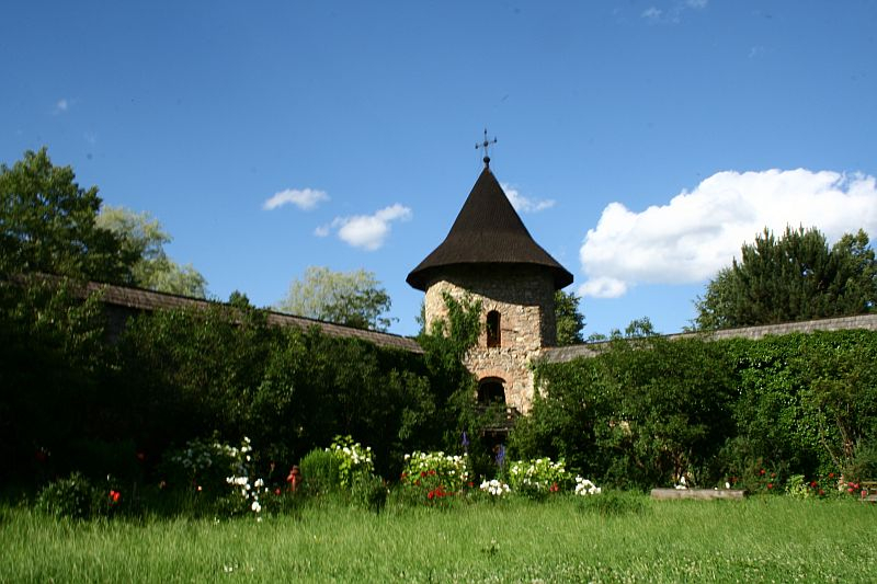 A tranquil garden with lush greenery and vibrant flowers under a clear blue sky, fronting an ancient stone tower with a conical roof, part of a historic walled compound in Romania.