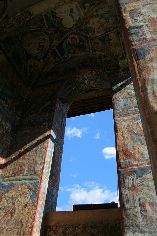 An ancient doorway adorned with intricate frescoes depicting religious scenes in Romania, opening to a bright blue sky, showcasing a blend of cultural history and natural beauty.