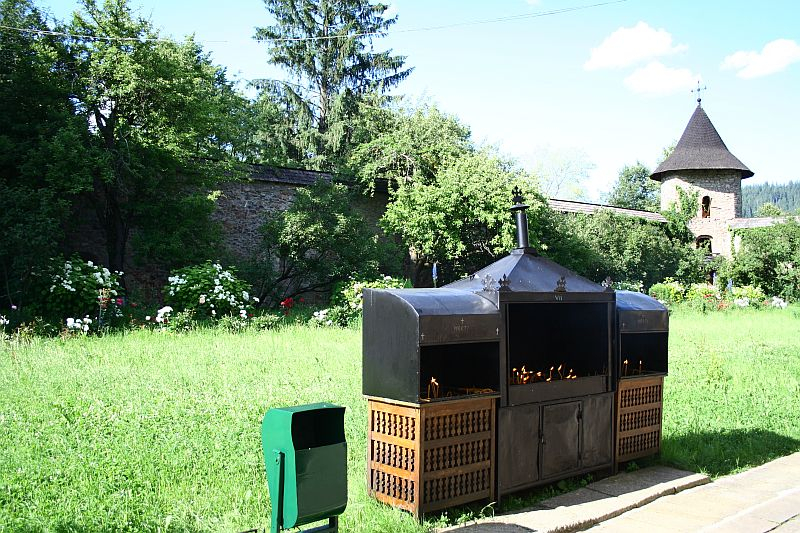 A large outdoor metal grill filled with lit charcoal is set in a verdant field with a green waste bin alongside, against a backdrop of an old stone wall and a medieval tower in Romania, surrounded by