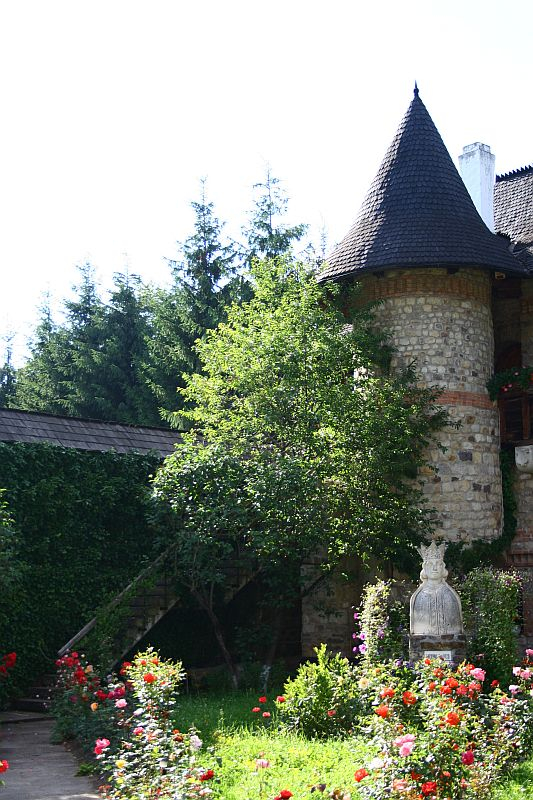 A quaint stone tower with a conical roof, reminiscent of Romania's countryside, is nestled amongst verdant trees and a vibrant garden of roses, creating a picturesque scene that exudes old-world charm.