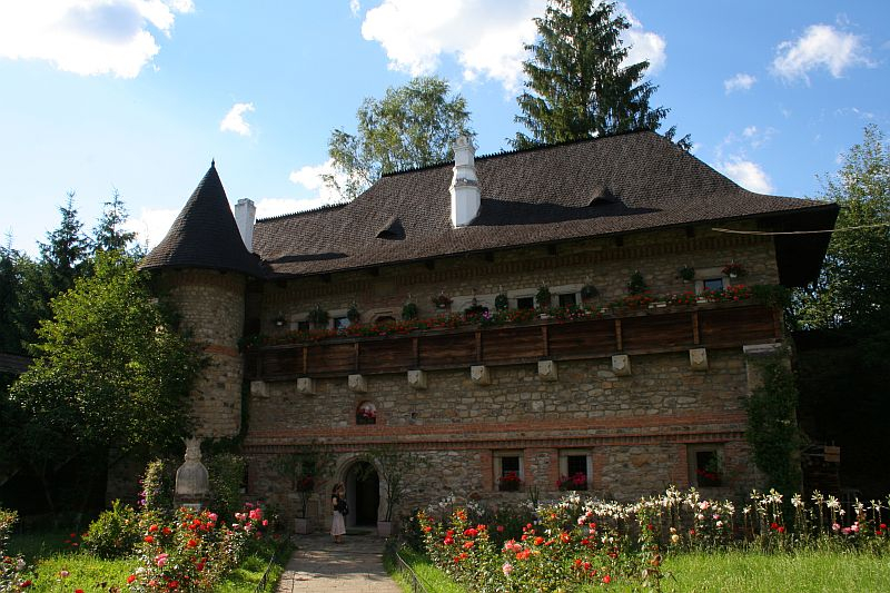A picturesque stone-built castle in Romania with a conical turret, featuring wooden balconies adorned with flowers, surrounded by lush greenery under a clear blue sky.