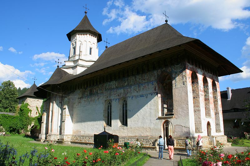 A historic church in Romania with a distinct stone bell tower and frescoed exterior walls, surrounded by lush greenery and flowering plants under a clear blue sky, while visitors approach its entrance.