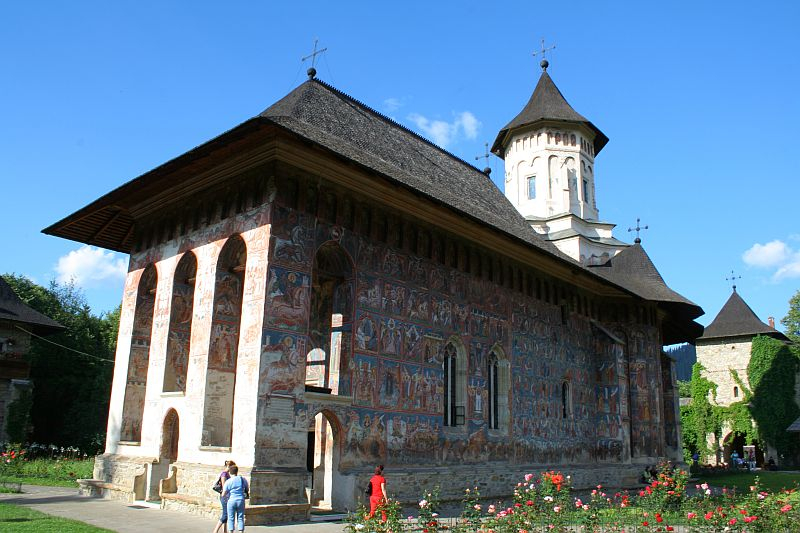 An ornate medieval monastery in Romania with vibrant frescoes on its exterior walls, under a clear blue sky, is admired by visitors strolling through its surrounding garden with blooming flowers.