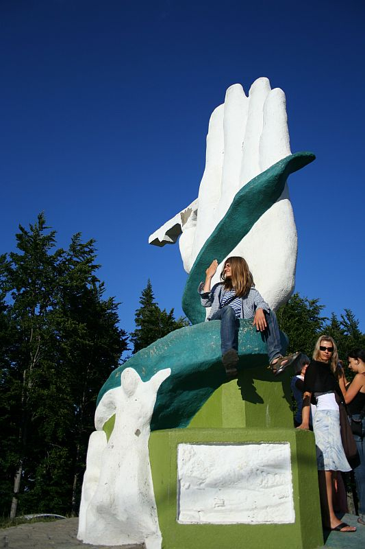 A person sits on a vibrant, oversized sculpture of a green and white hand in Romania, basking in the sunlight against a clear blue sky, surrounded by tall green trees.