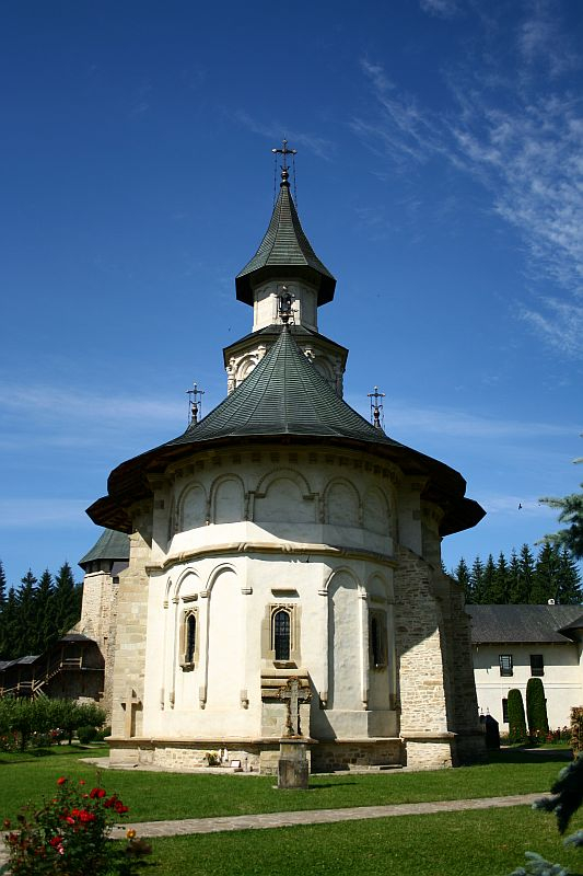 A historical church in Romania with a distinctive wood-clad spire and stone base stands gracefully under a clear blue sky, surrounded by manicured lawns and lush greenery, epitomizing architectural elegance