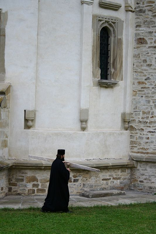 A solitary clergyman in traditional black vestments carries a blueprint while walking on the grass beside an old stone church in Romania, reflecting a blend of history and ongoing care or restoration.