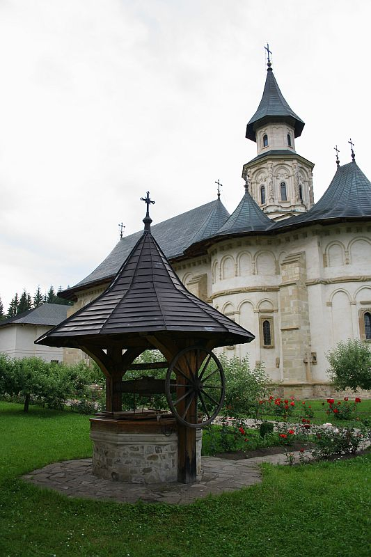 A traditional wooden well stands in the foreground of an ornate church with a prominent spire in Romania, surrounded by lush greenery and red flowers, evoking a serene, pastoral atmosphere.