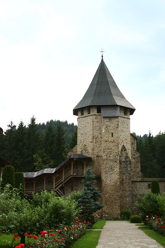An ancient stone tower with a conical roof stands guard beside a manicured pathway lined with vibrant flowers in Romania, with lush green trees under a cloudy sky in the background, evoking a sense of