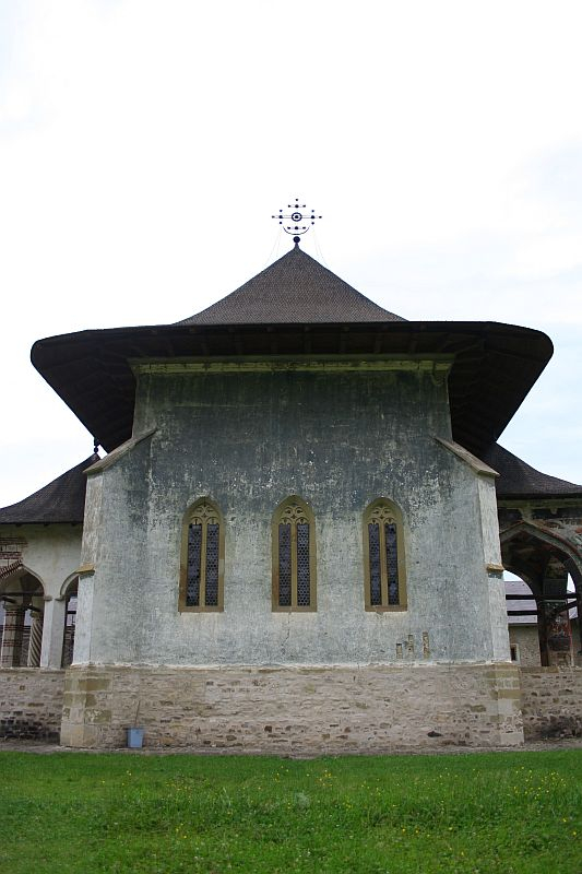 An ancient stone church with a large conical roof, featuring elongated windows and a cross atop, nestled in a serene grassy field in Romania.