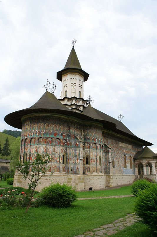 An historical monastery adorned with frescoes in Romania, nestled in a lush, green valley under a cloudy sky, showcases a traditional spired tower and a wrap-around porch with religious murals.