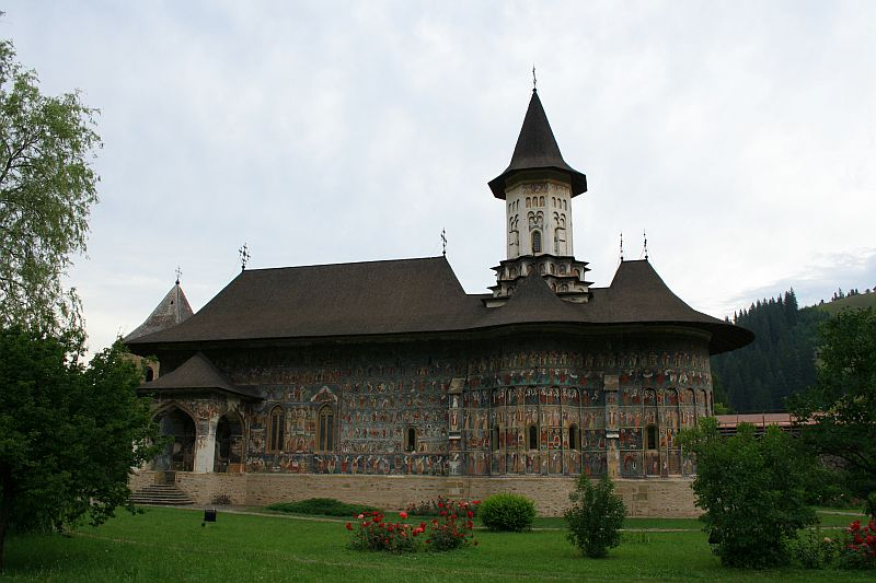 A traditional monastery in Romania with elaborate exterior frescoes and a prominent bell tower, surrounded by lush greenery under a cloudy sky.
