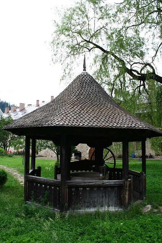 A traditional Romanian wooden well gazebo with a shingled roof, nestled in a lush green meadow with trees and a backdrop of white buildings with red roofs.