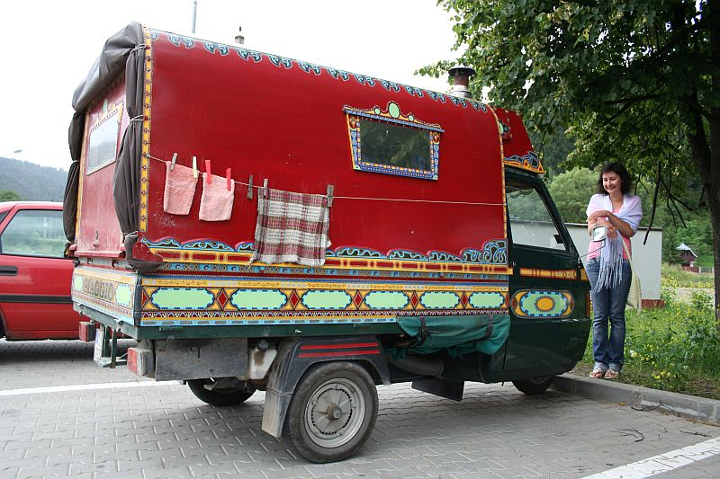 A woman stands beside a vibrantly decorated, red caravan with cultural Romanian patterns, parked in a roadside area, suggesting a glimpse into a nomadic or traveler lifestyle.
