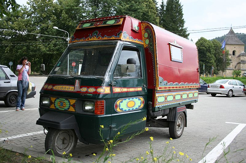 A woman talks on a mobile phone beside a colorfully painted, three-wheeled vehicle, resembling a decorated auto-rickshaw with an enclosed cargo area, parked on a sunny street in Romania with