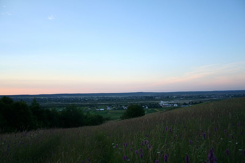 A serene dusk falls over the rural landscape of Romania, with the gentle slope of a flowering meadow in the foreground descending to a tapestry of fields and houses, all under the watchful gaze of