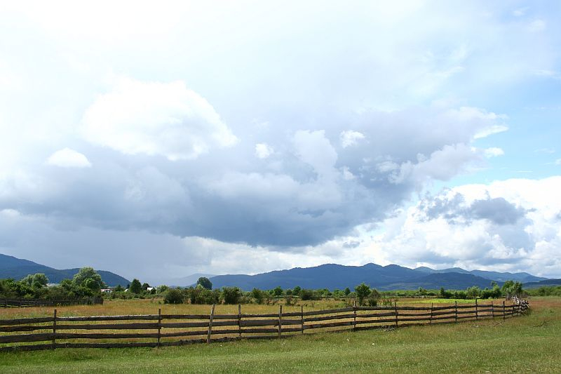 A serene rural landscape in Romania under a dramatic sky, with lush greenery, a rustic wooden fence leading the eye towards distant mountains, and heavy clouds suggesting an approaching storm.