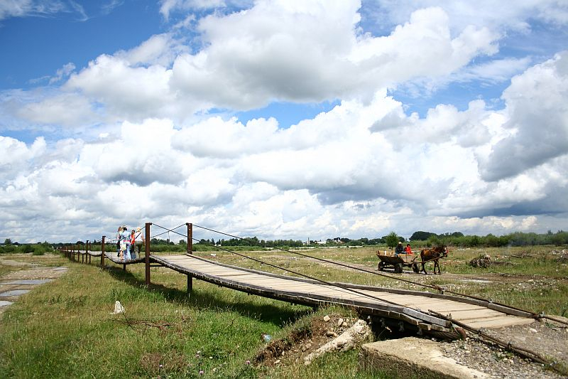 A rustic wooden bridge in Romania under a dynamic sky connects two stretches of land, with people walking across and a horse-drawn cart waiting nearby, showcasing rural life and transport.