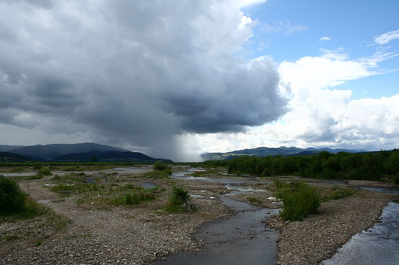 A vast, serene landscape in Romania with a meandering river amidst pebbled banks under a dramatic sky, where a localized rainstorm pours from a dark cloud onto distant hills, contrasting with the sun