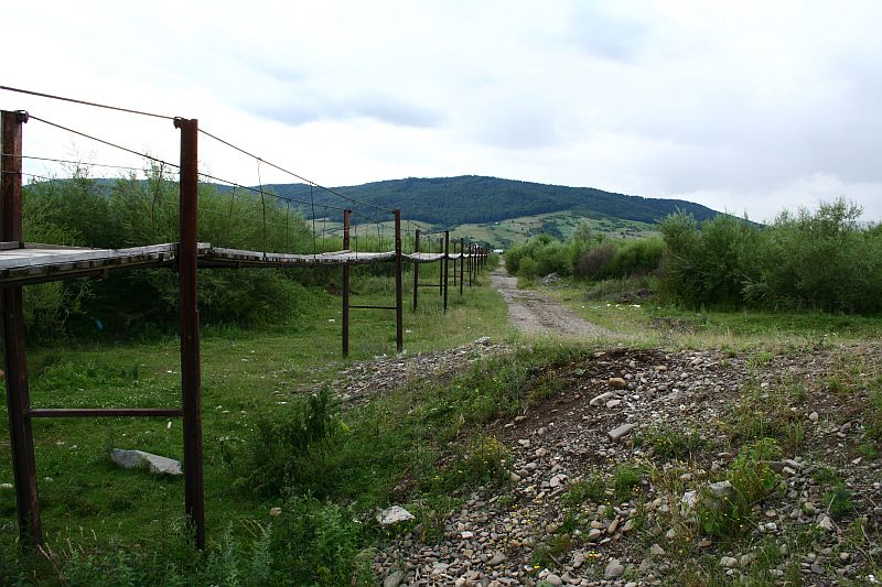 A rustic wooden footbridge with metal railings crosses over a dry rocky creek bed in Romania, leading towards a lush green valley with rolling hills under an overcast sky.