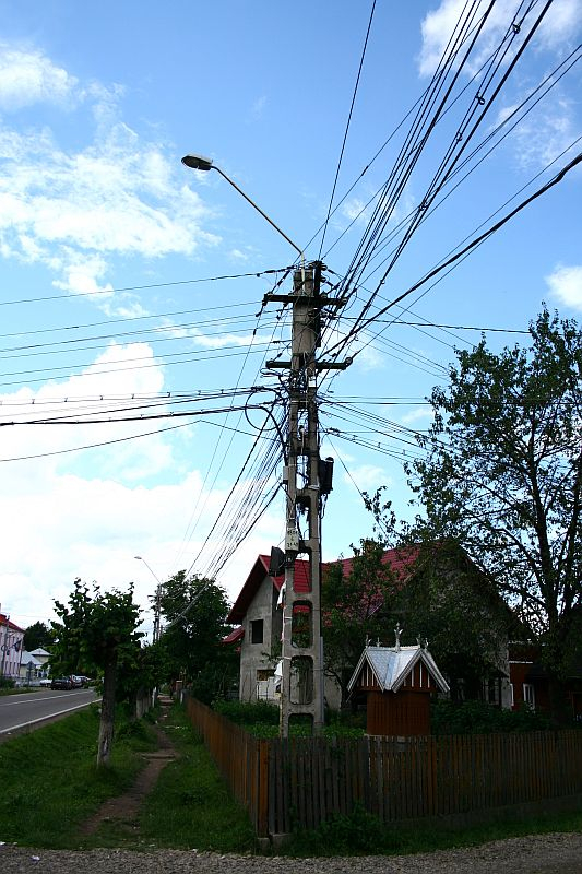 An electric pole stands at a rural street's edge in Romania, its arms entangled with a complex web of wires under a cloudy sky, flanked by greenery and quaint houses with picket fences