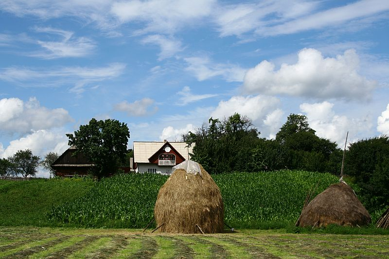 A picturesque rural landscape in Romania with a traditional house flanked by haystacks under a dynamic, cloud-streaked sky, emphasizing agricultural life.
